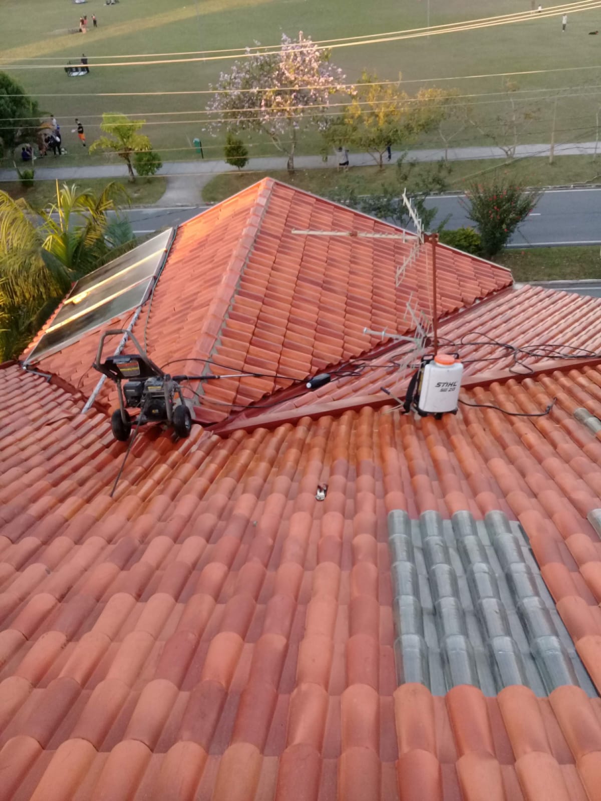 Telhado residencial com contraste evidente entre telhas já lavadas (laranja vibrante) e área ainda suja, com lavadora de alta pressão e pulverizador STIHL SG 20